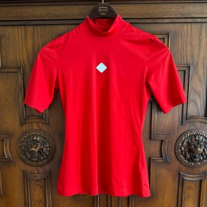 Röd golf polotröja J.Lindeberg - Snygg röd polotröja från J.Lindeberg med korta ärmar och diskret logga framtill. Tillverkad i mjuk och stretchig polyesterblandning, perfekt för golf eller sportiga dagar. Enkel och stilren design som sticker ut med sin starka färg.