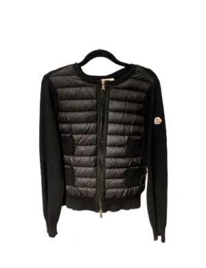Svart quiltad cardigan jacka Moncler - Svart cardigan jacka från Moncler med quiltad front och långa ärmar. Jackan har dragkedja hela vägen och Moncler-logga på ena ärmen. Materialmix med stickade partier och syntet på framsidan. Box från Moncler medföljer.
