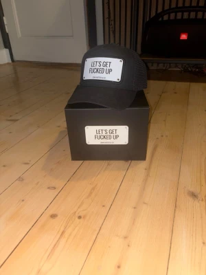 Svart keps John Hatter & Co - Säljer en svart keps från John Hatter & Co med texten 'LET'S GET FUCKED UP' på en vit patch framtill. Kepsen har mesh på sidorna och justerbar snapback. Perfekt för dig som vill sticka ut med en cool och edgy stil. Priset kan diskuteras .