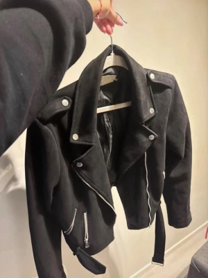 Svart croppad skinnjacka med bälte - Snygg svart mockajacka i bikerstil med silvriga dragkedjor, axelklaffar och bälte i midjan. Jackan har klassisk krage och detaljerade sömmar. Perfekt för dig som vill ha en edgy look.