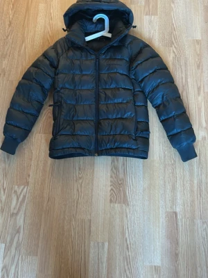 Svart pufferjacka med huva - Snygg svart pufferjacka med huva och dragkedja framtill. Jackan har quiltad design med breda kanaler och ribbade muddar vid ärmsluten. Perfekt för kalla dagar och enkel att matcha till olika outfits.