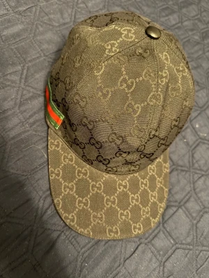 Gucci keps med GG-mönster - Säljer en snygg keps från Gucci i beige-brunt tyg med klassiskt GG-monogram över hela kepsen. På sidan finns en grön och röd rand som ger extra lyxig känsla. Kepsen har böjd skärm och metallknapp på toppen. Perfekt för dig som gillar streetstyle och designeraccessoarer.