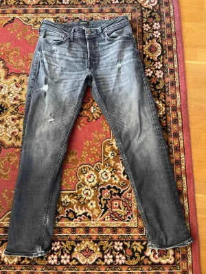 Grå slitna jeans från Jack & Jones - Säljer ett par grå jeans från Jack & Jones med slitningar och distressed detaljer på benen. Klassisk femficksmodell med knappgylf och raka ben. Jeansen har en cool tvättad look och passar perfekt till en avslappnad streetstyle. 31/30!
