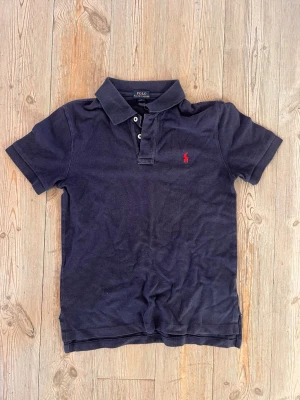 Marinblå pikétröja från Ralph Lauren - Klassisk marinblå pikétröja från Ralph Lauren med korta ärmar och krage. Tröjan har två knappar framtill och den ikoniska röda logotypen broderad på bröstet. Tillverkad i mjuk bomull som känns skön mot huden.