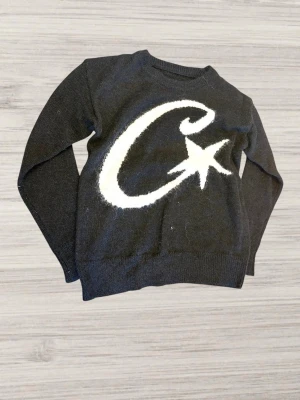 Svart stickad Corteiz tröja - Cool svart stickad tröja med ett stort vitt C och en stjärna på bröstet. Tröjan har rund halsringning och långa ärmar. Perfekt för dig som gillar grafiska tryck och vill sticka ut lite extra. Skriv till mig vid funderingar.