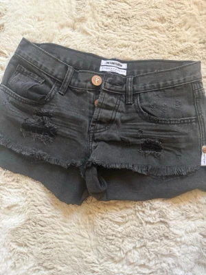 Svarta denimshorts med slitningar - Säljer ett par svarta denimshorts från One Teaspoon med råa kanter och slitna detaljer framtill. Klassisk femficksmodell med knappgylf och låg midja. Perfekta för en avslappnad och trendig sommarstil.