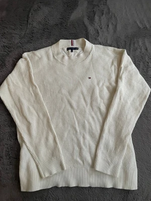 Vit stickad tröja Tommy Hilfiger - Klassisk vit stickad tröja från Tommy Hilfiger med rund halsringning och diskret logga broderad på bröstet. Tröjan har ribbade muddar vid ärmslut och nederkant samt en mjuk, strukturerad stickning. Perfekt för en stilren och enkel look.