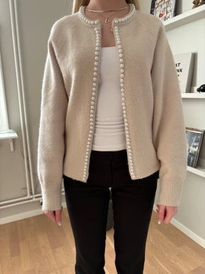 Beige kofta med pärldetaljer - Snygg beige kofta med rund hals och pärldetaljer längs hela öppningen och halsringningen. Koftan har långa ärmar och ribbade muddar vid ärmslut och nederkant. Perfekt för dig som gillar en stilren men ändå unik look. Men endast en knapp upptill. 