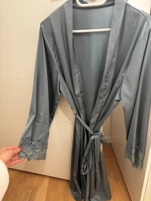 Ljusblå satinmorgonrock med spets - Sexig ljusblå morgonrock i satin med knytband i midjan och breda ärmar. Ärmsluten har fina spetsdetaljer som ger en elegant touch. Aldrig använd. Tvättat den. Fått i present så ingen aning vart den är köpt. 