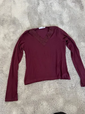 Vinröd långärmad topp Gina Tricot - Snygg vinröd långärmad topp från Gina Tricot med spetsdetaljer runt v-ringningen. Perfekt för dig som gillar en enkel men ändå elegant stil. Tillverkad i mjukt material som känns skönt mot huden.