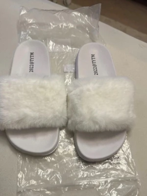 Vita fluffiga tofflor med fuskpäls - Säljer ett par vita tofflor från WALLY med bred rem i mjuk fuskpäls. Sulan är i plast och har struktur undertill för bättre grepp. Perfekta för att chilla hemma eller ge din outfit en cozy vibe.