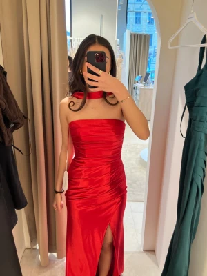 Röd långklänning med slits - Elegant röd långklänning i glansigt material med axelbandslös design och matchande choker. Klänningen har en smickrande drapering som framhäver figuren samt en hög slits framtill som ger en stilren och feminin look.  Klänningen är helt oanvänd med prislappen kvar och köpt från Dressroomstudio. Perfekt för bal, fest eller andra speciella tillfällen där du vill sticka ut.  Storlek 34 och i nyskick.