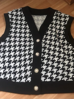 Svartvit rutig väst med knappar - Snygg svartvit väst med klassiskt rutigt mönster och dekorativa knappar framtill. Västen har v-ringad hals.
