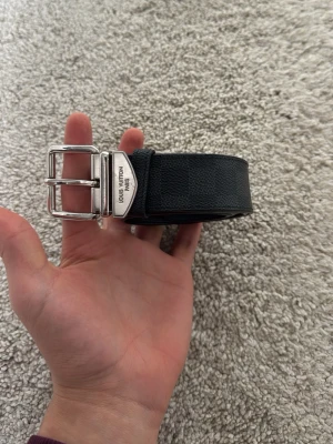 Svart rutigt bälte Louis Vuitton - Snyggt svart bälte från Louis Vuitton med klassiskt rutigt mönster och silverfärgat spänne. Bältet är gjort i skinn och har en stilren design som passar till många olika outfits. Bältet är 100% äkta då den är köpt i Louis Vuitton affären. Pris kan diskuteras vid snabb affär 