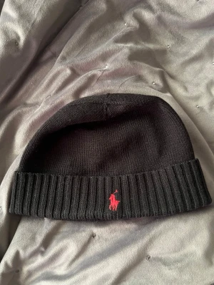 Svart mössa från Polo Ralph Lauren - Säljer en svart ribbstickad mössa från Polo Ralph Lauren med den klassiska röda loggan broderad framtill. Mössan har uppvikt kant och är tillverkad i mjukt stickat material som håller dig varm under kalla dagar. Köpt från kidsbrandstore