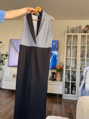 Svart och vit ärmlös jumpsuit - Snygg ärmlös jumpsuit med svart och vit hundtandsmönstrad överdel och svart nedtill. Djup V-ringning med bred krage ger en cool och stilren look. Perfekt för dig som vill sticka ut lite extra. Materialet känns mjukt och följsamt.