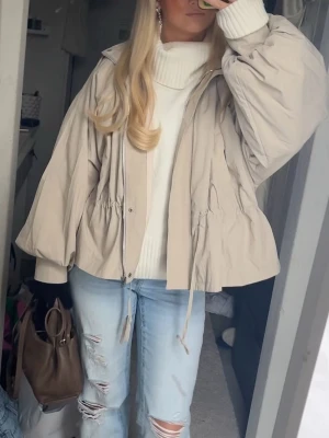 Jacka med snörning - Snygg beige jacka med ballongärmar och dragsko i midjan för en justerbar passform. Jackan har dragkedja framtill och en avslappnad, loose siluett. Perfekt för lager på lager och enkel att styla till olika outfits. Tunn i materialet så får plats med det mesta 
