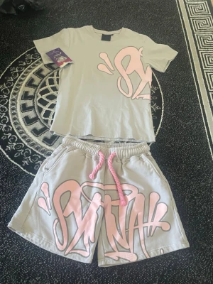 Grå shorts med rosa graffiti-tryck - Säljer ett par grå shorts med stort rosa graffiti-inspirerat tryck framtill och rosa snörning i midjan. Shortsen har avslappnad passform och sidofickor. Perfekta för dig som gillar streetwear och vill sticka ut.