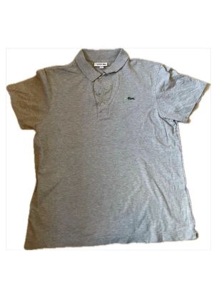 Grå pikétröja från Lacoste - Klassisk grå pikétröja från Lacoste med krage, knappar framtill och den ikoniska krokodilloggan broderad på bröstet. Tillverkad i mjuk bomull och har korta ärmar. Perfekt för en clean och sportig look.