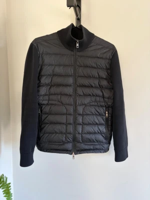 Svart cardigan och tibb väst från Moncler - Snygg svart dunväst från Moncler med hög krage och quiltad design. Västen har två dragkedjeförsedda fickor och Moncler-logga på bröstet. Perfekt för lager-på-lager och ger en sportig, trendig look.