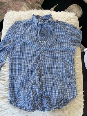 Blårutig skjorta från Polo Ralph Lauren - Klassisk blårutigu skjorta från Polo Ralph Lauren med button-down krage och broderad logga på bröstet. Skjortan har långa ärmar, vita knappar och är tillverkad i mjuk bomull. Perfekt för en avslappnad men ändå stilren look.