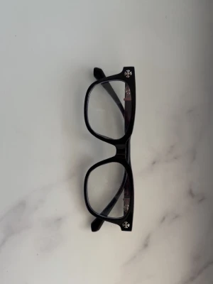 Chrome heart glasögon  - Stilrena svarta solglasögon med markerad båge och fyrkantig form. På bågens hörn och skalmar finns snygga silverfärgade korsdetaljer ( chrome heart)  som ger en unik touch. Perfekta för dig som vill ha en cool accessoar med edge.