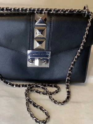 Svart axelväska Michael Kors nitar - Snygg svart axelväska från Michael Kors i läder med silverfärgade metalldetaljer och kedjerem. Väskan har coola pyramidformade nitar framtill och ett stilrent lås. Perfekt för dig som gillar en edgy och trendig look.