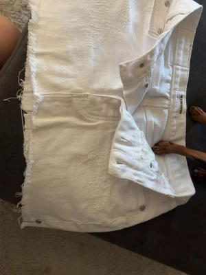 Vit jeanskjol med rå kant från one teaspoon - Säljer en vit jeanskjol från one teaspoon i storlek w24 med rå, fransig kant nertill och slitna detaljer framtill. Kjolen har klassisk femficksdesign, knappgylf och är i ett kraftigt bomullstyg. Perfekt för dig som gillar en avslappnad och trendig stil.