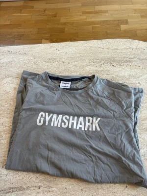 Grå Gymshark t-shirt med tryck - Snygg grå t-shirt från Gymshark med stort vitt logotryck på bröstet. Klassisk rund halsringning och mjukt material som känns skönt mot huden. Perfekt för dig som gillar sportiga och stilrena plagg.