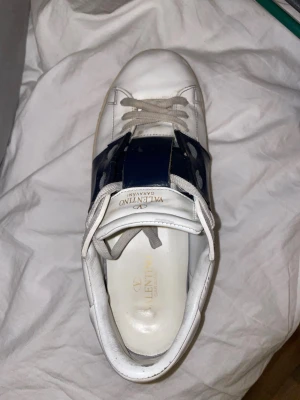 Valentino vit/blå sneaker  - Snygg vit sneaker från Valentino med bred blå rem över snörningen och diskret logga på plösen. Skon har klassisk låg profil och är tillverkad i läder med gummisula. Perfekt för dig som gillar exklusiva och stilrena sneakers. Äkta skriv om ni vill ha mer bilder och pris kan diskuteras med snabb affär 