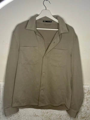 Beige overshirt från Zara - Stilren beige overshirt från Zara med två bröstfickor och klassisk krage. Overshirten har knäppning framtill och är tillverkad i ett mjukt, strukturerat material. Perfekt att bära öppen eller stängd för en avslappnad look.