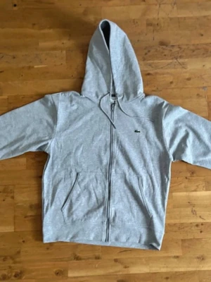 Gray Lacoste zip hoodie - Snygg grå zip hoodie från Lacoste med klassisk krokodillogga på bröstet. Hoodien har dragkedja, två fickor fram och justerbar huva med snören. Tillverkad i mjukt bomullsmaterial som känns skönt mot huden. Perfekt för en avslappnad och stilren look.