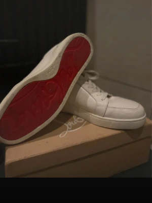 Vita Louboutin sneakers med röd sula - Säljer ett par vita sneakers från Christian Louboutin med den ikoniska röda sulan. Skorna har en clean och stilren design i läder, lågt skaft och klassisk snörning. Perfekta för dig som vill ha något exklusivt och trendigt i garderoben.