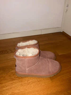 Rosa UGG boots med fårskinnsfoder - Mysiga rosaaktiga UGG boots i mocka med fluffigt vitt fårskinnsfoder. De har en låg modell med rund tå och grov sula i gummi. Perfekta för kalla dagar när du vill hålla fötterna varma och snygga.