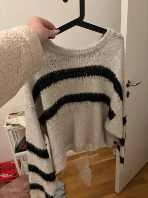 Grå stickad tröja med svarta ränder - En oversized stickad tröja i ljusgrått med breda svarta horisontella ränder över bröstet och ärmarna. Tröjan har rund halsringning och långa ärmar. Perfekt för en avslappnad och trendig look.