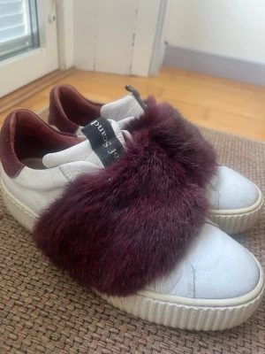Sneakers med päls - Unika vita sneakers från Pura López med bred vinröd fuskpälsdetalj över vristen och vinröda detaljer vid hälen. Skorna har en chunky sula och svart logotypband på plösen. Tillverkade i läder och fuskpäls för en trendig look. Använda fåtal gånger men inte alls smutsiga eller repiga. Storlek 38 men passar lite mindre och större. Mörk Lila/Vinrött