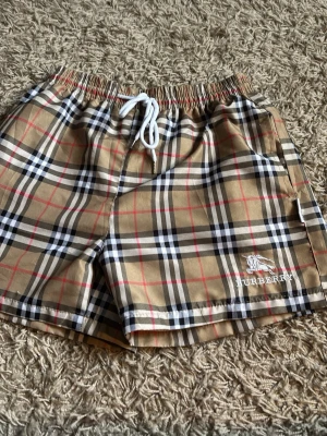 Rutig Burberry shorts med snörning - Säljer ett par klassiska Burberry shorts i beige, svart, vit och röd rutig design. Shortsen har elastisk midja med vit snörning och Burberry-logga broderad på benet. Dom är storlek M men sitter som S. Dom är helt oanvända 