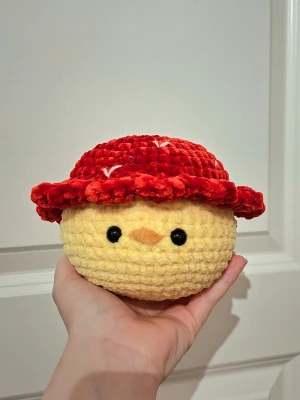 Gul virkad kyckling med röd hatt - Söt handgjord plushie som föreställer en gul kyckling med röd jordgubbsinspirerad hatt gjort på chenillegarn. Den har även sitt egna namn som sitter på produkten vid köp. Färgerna på bilden kan vara lite annorlunda i verkligheten. Eftersom produkten innehåller säkerhetsögon rekommenderas den inte för barn under 3 år. 