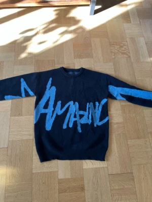 Svart stickad tröja med blå text - Cool svart stickad tröja med stor blå text i graffiti-stil över hela framsidan och ärmarna. Rund halsringning och långa ärmar. Perfekt för dig som vill sticka ut med en unik och trendig look.