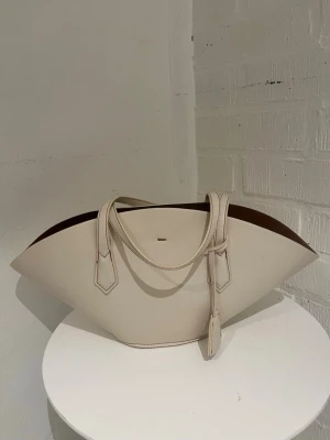 Vit handväska från Zara - Stilren handväska från Zara i vitt konstläder med unik, bred form och två handtag. Väskan har en minimalistisk design och insidan är fodrad i ljust tyg. Perfekt för dig som gillar trendiga accessoarer med modern känsla.