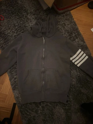 Thom browne - Mysig grå hoodie med dragkedja och huva. Ena ärmen har tre vita ränder och på ryggen finns ett stort segelbåtsmotiv i vitt, svart och rött. Två fickor framtill och ribbade muddar. Perfekt för en avslappnad och sportig stil.
