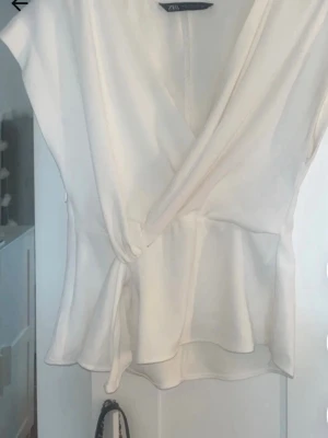 Vit omlottopp från Zara - Snygg vit omlottopp från Zara med knytband i midjan och lätt v-ringad hals. Toppen har korta ärmar och ett mjukt, följsamt tyg som ger en avslappnad men chic look. Perfekt att styla med både jeans och kjol. Sömnen har lossnat på ena armén längst ut men går att fixa lätt.