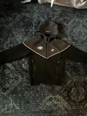 Svart Nike teech - Säljer ett svart loungewear set från Nike med matchande hoodie och byxor. Setet har vita detaljer och loggor på både tröja och byxa samt dragkedjor på fickorna. Perfekt för chill dagar hemma eller när du vill vara bekväm men ändå se snygg ut.