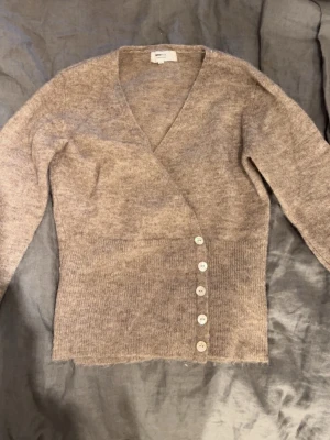 Beige kofta - Mysig beige omlottkofta med långa ärmar och fem vita knappar framtill. Koftan har en djup v-ringning och är stickad i ett mjukt material. Knappt använd och är slutsåld på Gina’s hemsida. Nypris 460kr