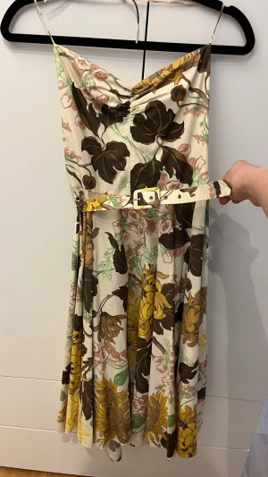 Blommig klänning - Säljer en blommig klänning med smala axelband och vackert fall. Klänningen har ett matchande bälte i midjan och ett färgstarkt mönster i brunt, gult, grönt och beige på ljus botten. Perfekt för sommarens alla tillfällen!