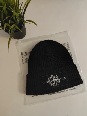 Svart ribbstickad mössa Stone Island - Svart ribbstickad mössa från Stone Island med uppvikt kant och den klassiska logotypen framtill. Snygg och enkel design som passar perfekt till kalla dagar. Materialet är mjukt och värmande, perfekt för höst och vinter.