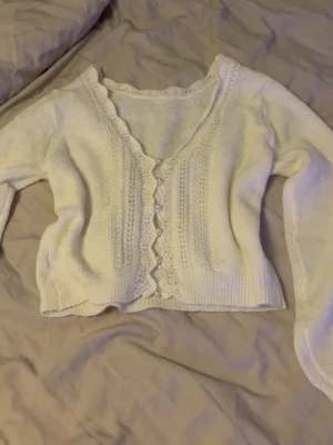 Vit söt kofta med pärlknappar - Supersöt vit kofta med v-ringning och dekorativa pärlknappar framtill. Koftan har ett vågigt kantmönster längs knäppningen och är stickad i ett mjukt material. Långa ärmar och ribbade muddar ger en mysig känsla.