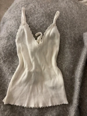 Vit ribbad linnetopp med spetskant - Söt vit ribbstickat linne med smala axelband och v-ringning. Toppen har fina kanter och knytning i ryggen för extra detalj. Ger en illusion av en större byst. Perfekt för lager på lager eller till sommaren.