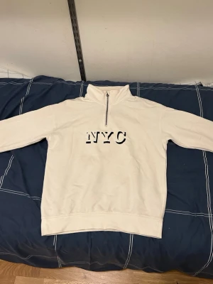 NYC hoodie  - This cream-colored quarter-zip sweatshirt features a relaxed, drop-shoulder fit and a high collar with a silver metallic zipper. The chest is detailed with a bold "NYC" graphic in a black, distressed serif font. Constructed with ribbed cuffs and a ribbed hem, this piece offers a clean, streetwear-inspired aesthetic.