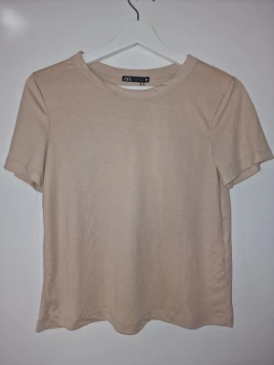 Beige basic t-shirt från Zara - En stilren beige t-shirt från Zara med enkel design som gör den mångsidig och lätt att matcha med olika underdelar. Passar bra för en avslappnad vardagslook.